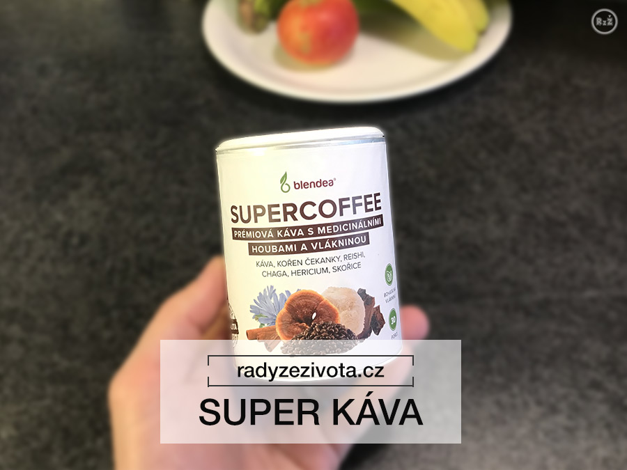 Fotografie instantní kávy SUPERCOFFEE v papírové nádobě držena v ruce v pozadí kuchyňská linka a talíř s ovocem